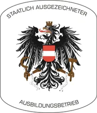 Emblem eines Ausbildungsbetriebs mit einem Doppeladler, der Ketten, Hammer und Sichel hält, mit der österreichischen Flagge in der Mitte. Der Name des Instituts ist in Deutsch geschrieben.