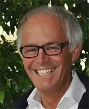 Ein Mann mit weißen Haaren, der eine Brille und ein weißes Hemd trägt, lächelt und steht vor einer grünen Pflanze.