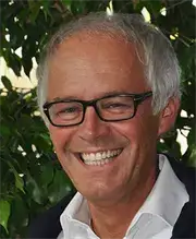 Ein Mann mit weißen Haaren, der eine Brille und ein weißes Hemd trägt, lächelt und steht vor einer grünen Pflanze.