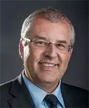 Ein Mann in Anzug und Krawatte, mit Brille, lächelt fröhlich in die Kamera. Der Hintergrund ist dunkel.