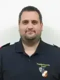 Ein Mann in einer schwarzen Polizeiuniform steht lächelnd vor einer weißen Wand. Das Abzeichen auf seinem Shirt lautet 'Polizist'.