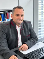 Ein Mann in einem grauen Jackett mit rotem Rand sitzt an einem Schreibtisch und schreibt auf einem Stück Papier mit einem Stift.