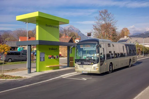 Ein Bus ist an einem grünen Busbahnhof geparkt, mit Bergen im Hintergrund. Der Bus hat ein Schild, das Postbus sagt.