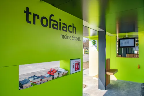 Ein grüner Kiosk mit 'trofaiaich meine stadt' darauf. Eine Defibrillatorbox ist an der Wand montiert. Bücher sind auf dem Regal angeordnet. Eine Holzbank ist auf der rechten Seite.