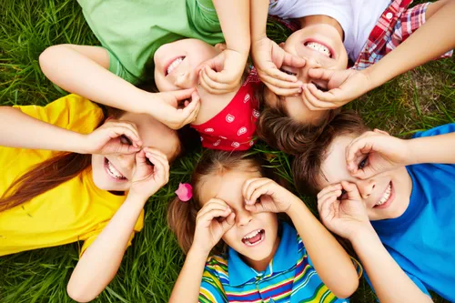 Gruppe junger Kinder liegt auf dem Gras, lächelt und posiert mit den Fingern an den Augen. Sie sind in einer kreisförmigen Formation, wobei jedes Kind zur Kamera schaut.