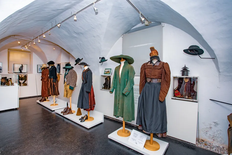 Ein Museum zeigt historische Kleidung und Hüte auf Mannequins. Verschiedene Kleidungsstücke sind in einer Reihe angeordnet mit Etiketten. Weiße Wände und Deckenleuchten verbessern die Sichtbarkeit.