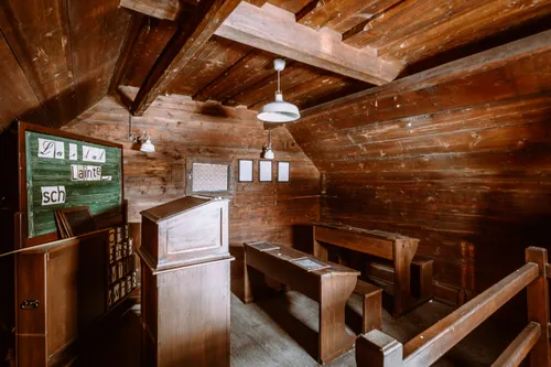 Ein altes Klassenzimmer mit Holzwänden, Bänken und einer Tafel. Zwei Lampen hängen von der Decke.