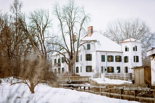 Ein großes weißes Gebäude mit grünen Fensterläden und einem Schornstein, mit Schnee bedeckt, steht in einer winterlichen Landschaft mit Bäumen und einem Holzzaun.