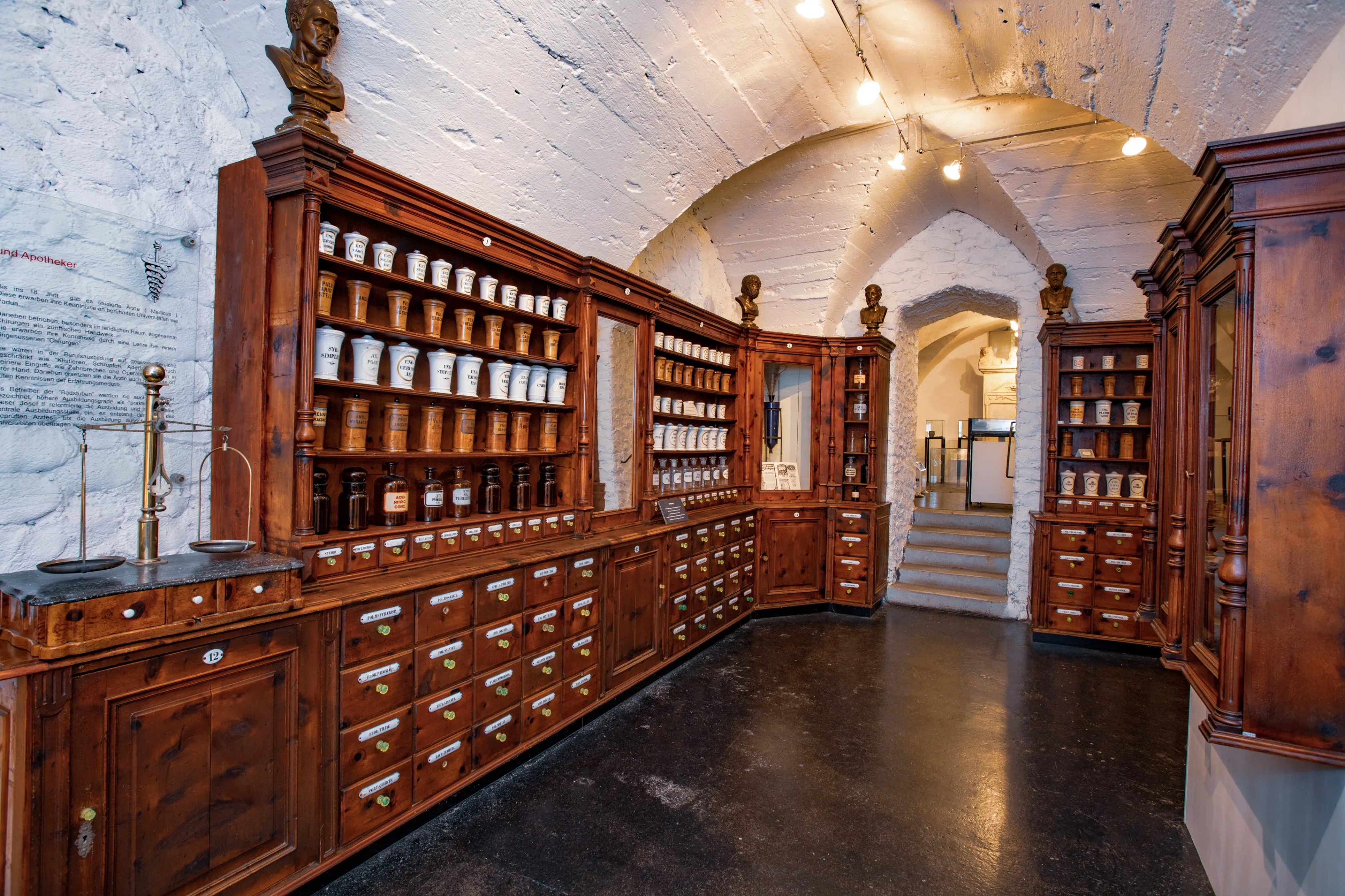 Eine alte Apotheke mit Holzregalen voller Gläser und Flaschen, beleuchtet von Lampen an der Decke, mit Büsten über den Regalen und einer Treppe, die nach rechts führt.