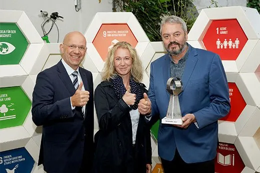 Drei Personen, zwei Männer und eine Frau, posieren mit einem Pokal vor einer sechseckigen Struktur. Sie lächeln und geben Daumen hoch.