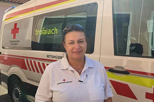 Eine Frau in einem weißen Shirt mit rotem Kreuz steht vor einem Krankenwagen. Der Krankenwagen hat rote und gelbe Markierungen.