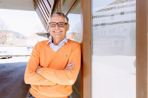 Ein Mann in einem orangefarbenen Pullover steht mit verschränkten Armen vor einem Gebäude. Er lächelt in die Kamera.