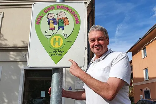 Ein Mann steht neben einem weißen Schild mit einem grünen Logo, zeigt darauf und deutet darauf hin, mit Gebäuden und Autos im Hintergrund.