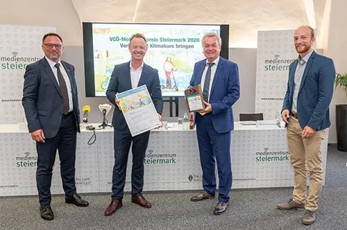 Vier Männer in formeller Kleidung stehen vor einem Podium mit einer großen Leinwand im Hintergrund, alle lächeln und halten Auszeichnungen.