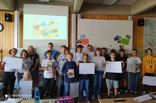 Ein Klassenzimmer voller Schüler, die Papiere halten, vor einer Tafel mit einem Projektionsbild darüber. Der Raum ist mit Holz verkleidet, an der Wand hängt eine Uhr.