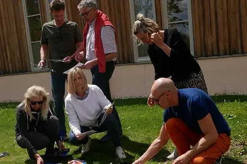Eine Gruppe von Menschen steht und kniet auf einem Rasen und liest Papiere. Sie befinden sich vor einem Gebäude mit Glasfenstern.