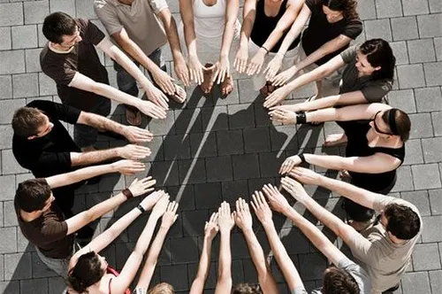 Eine Gruppe von Menschen steht im Kreis und hält sich an den Händen. Sie sind lässig gekleidet, einige tragen eine Brille. Der Boden ist gepflastert und sie werfen Schatten.