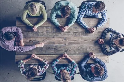 Eine Gruppe von Menschen sitzt um einen Holztisch herum, vielleicht in einer Besprechung. Einige lehnen sich auf den Tisch, während andere nach unten schauen.