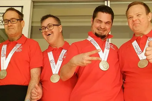 Vier Männer in roten Poloshirts und mit Brillen tragen Medaillen um den Hals und posieren für ein Foto. Sie alle haben fröhliche Gesichtsausdrücke.