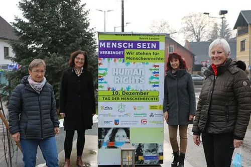 Vier Frauen stehen vor einem Banner auf einem Bürgersteig. Sie lächeln und scheinen für ein Foto zu posieren. Das Banner hat ein Menschenrechtsthema.