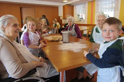 Eine Gruppe älterer Frauen und Kinder sitzt an einem Tisch. Sie tragen Schürzen und scheinen zu backen.