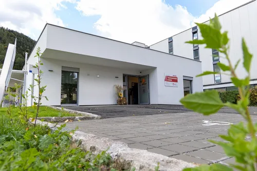 Ein modernes weißes Gebäude mit Glasfassade und offenen Eingangstüren, umgeben von Grün und einer gepflasterten Auffahrt.
