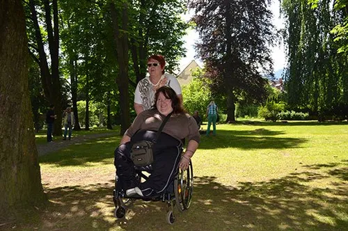 Zwei Frauen sind in einem Park. Eine Frau ist im Rollstuhl und eine andere steht hinter ihr. Sie sind von Grün umgeben.