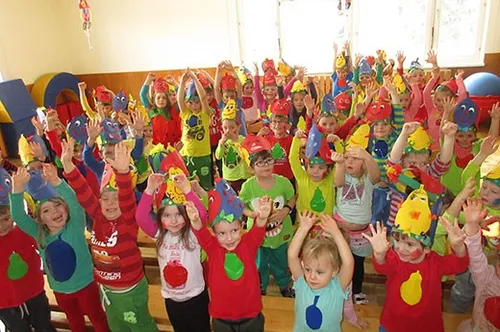Ein Klassenzimmer voller Kinder, alle in bunten Kostümen gekleidet, heben ihre Hände und lächeln. Sie tragen Mützen in Form von Obst und Gemüse. Einige von ihnen halten Requisiten. Dahinter steht ein gelbes Objekt, möglicherweise ein Ball.
