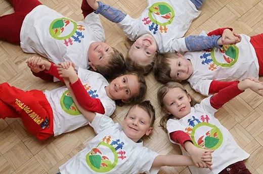 Sieben junge Kinder liegen auf einem Holzboden und tragen passende weiße T-Shirts mit einem farbenfrohen Cartoon-Charakter-Design. Sie scheinen für ein Foto zu posieren, mit den Händen ineinander verschränkt. Das Kind auf der rechten Seite trägt rote Hosen.