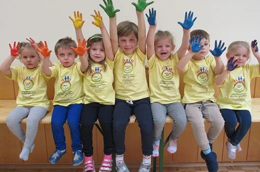 Eine Gruppe von Kindern mit gelben T-Shirts und Logos und bunten Handabdrücken auf einem Stuhl sitzen. Sie tragen verschiedenfarbige Hosen und Schuhe.