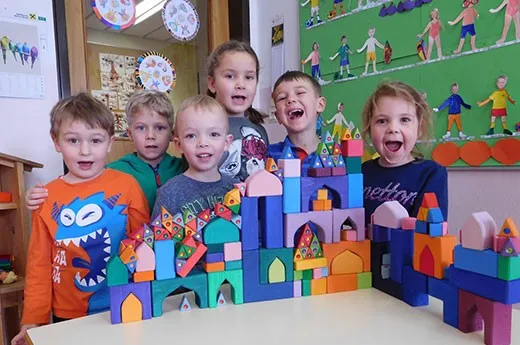 Fünf Kinder stehen um einen Tisch herum, der mit bunten Bauklötzen bedeckt ist, lächeln und posieren für die Kamera.