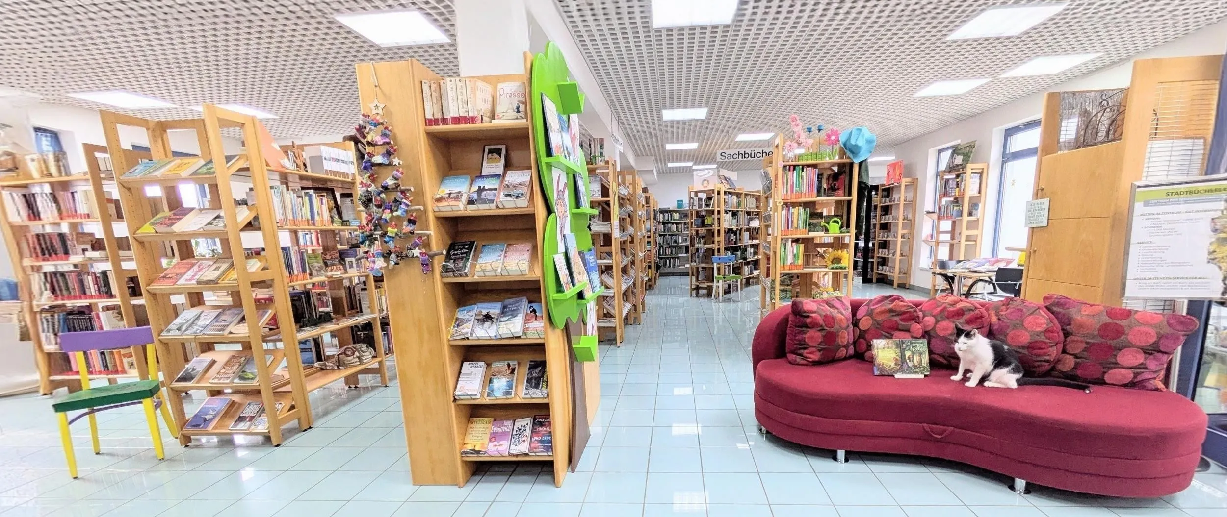 Eine Buchhandlung mit Holzregalen, gefüllt mit Büchern und Dekorationsartikeln. Auf der rechten Seite befindet sich ein gepolsterter Sessel mit Kissen. Die Decke hat weiße Paneele und Beleuchtung.