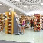 Eine Frau benutzt einen Geldautomaten in einer Bibliothek. Mehrere Leute stehen herum und stöbern in Büchern auf den Regalen. Die Bibliothek hat ein modernes Design mit weißen Fliesen und Holzregalen.