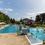 Ein Freibad mit Schwimmenden und einer Rutsche. Der Pool ist von Bäumen umgeben, im Hintergrund Berge.
