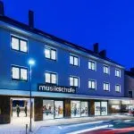 Ein Musikschulgebäude mit Glasfenstern, das bei Nacht beleuchtet ist. Es trägt ein Schild mit der Aufschrift 'musikschule' und mehrere Personen stehen davor.