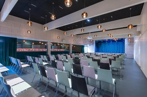 Ein Konferenzraum mit Reihen von Stühlen, die auf eine Bühne mit blauem Vorhang ausgerichtet sind. Die Decke ist mit mehreren hängenden Lampen ausgestattet. Die Wände haben ein gemustertes Design und der Boden ist grau.