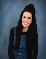 Eine Frau mit langen schwarzen Haaren lächelt und trägt eine Jeans-Latzhose mit einem schwarzen Langarmshirt darunter. Sie steht vor einem blauen Hintergrund.