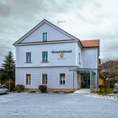 Ein weißes Gebäude mit rotem Rand und einem roten Dach zeigt den Namen 'Gemeindeamt'. Es ist von einem Kiesplatz mit Pflanzen und Bäumen umgeben. Ein Auto ist links geparkt.