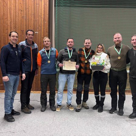 Eine Gruppe von Menschen in Winterjacken und Medaillen posiert für ein Foto in einer Sportanlage mit einer Holzwand und einem grünen Netz im Hintergrund.