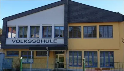 Ein gelbes Gebäude mit schwarzem Dach und mehreren Fenstern. Über dem Eingang steht ein Schild mit der Aufschrift 'Volksschule'.