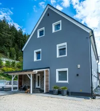 Ein graues Haus mit einer Ziegelmauer, weißen Fenstern und einem Hund, der vor dem Eingang sitzt.
