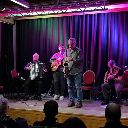 Vier Musiker auf der Bühne, einer spielt Saxophon, ein anderer Gitarre, und zwei spielen Akkordeon. Ein Publikum sitzt davor.