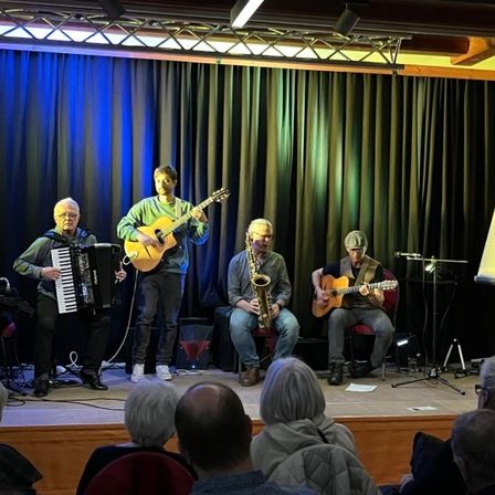 Vier Musiker spielen auf der Bühne unter blauen Lichtern vor einem Publikum. Die Musiker spielen Gitarre, Saxophon, Akkordeon und Schlagzeug.