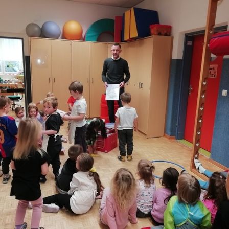 Ein Mann steht in einem Spielzimmer mit einer Gruppe von Kindern, einige sitzen und andere stehen herum. Bunte Spielzeuge und Bälle sind verstreut.