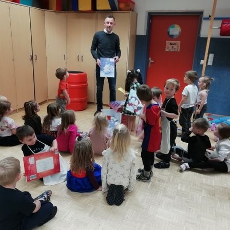 Eine Gruppe von Kindern und einem Mann versammelt sich in einem Raum. Der Mann hält ein Buch und die Kinder lauschen aufmerksam.