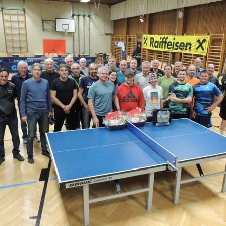 Eine Gruppe von Menschen posiert für ein Foto vor einem Tischtennistisch. Sie stehen in einer Turnhalle.