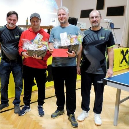 Vier Männer stehen in einer Sporthalle zusammen und halten Geschenkkörbe. Hinter ihnen hängt ein gelber Banner und ein Tischtennistisch ist zu sehen.