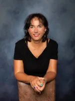 Eine Frau mit kurzen Haaren lächelt und sitzt auf einem braunen Stuhl, trägt ein schwarzes V-Neck-Shirt. Ihre Hände sind vor ihr verschränkt.