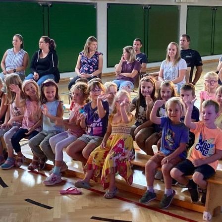 Kinder und Erwachsene sitzen auf Holzbänken in einer Turnhalle, schauen nach vorne und winken. Einige tragen Brillen und Turnschuhe. Eine Frau hält eine Wasserflasche.