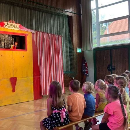 Eine Gruppe von Kindern sieht sich ein Puppentheater auf der Bühne an, mit einem rot-weißen Vorhang und einer gelben Struktur mit dem Schriftzug 'Stringle Puppentheater'.